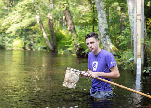 A student in a 51小黄车 shirt stands in the Blackstone River with a bug catching net